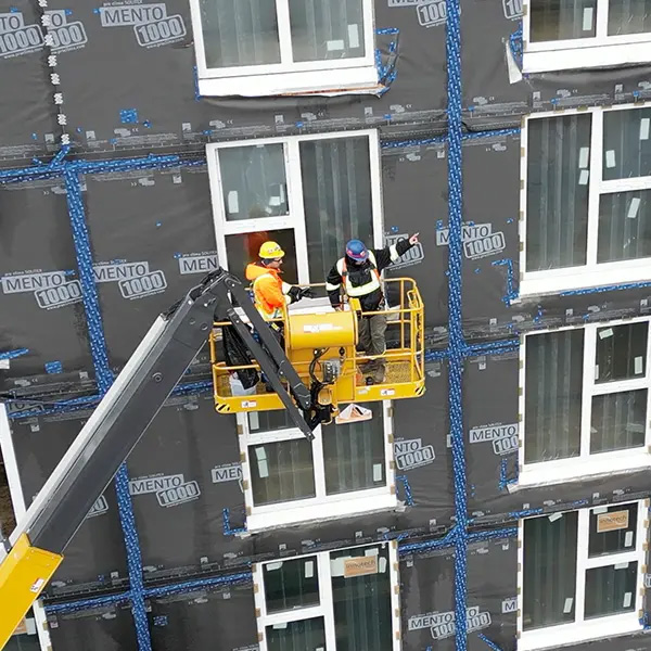 Two workers in a lift installing sheathing on a building with white-framed windows.