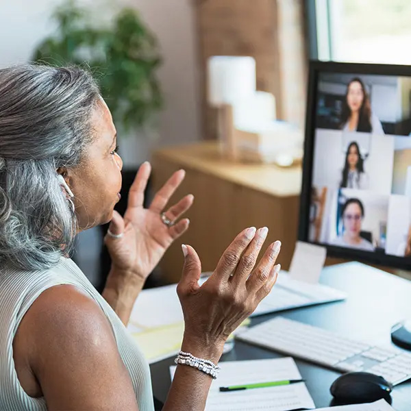 Woman sitting at a desk during a video conference, with blurred faces on the monitor and various items like papers, keyboard, and mouse on the desk.