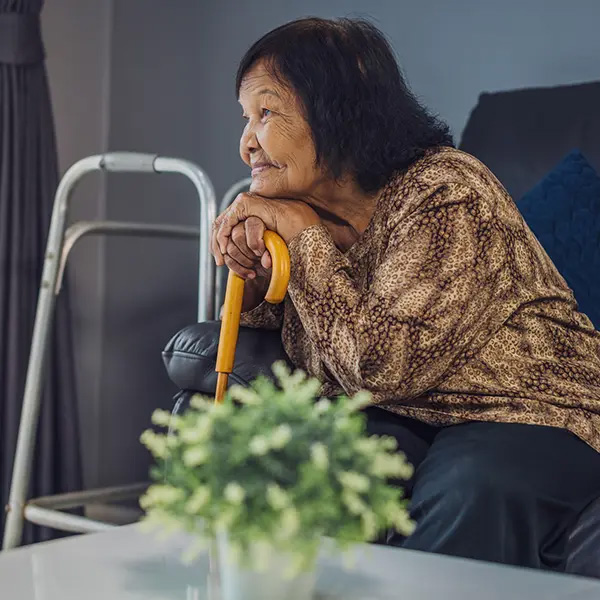 Elderly person sitting on a couch, holding a yellow cane with both hands, with a walker nearby and a potted plant in the foreground.