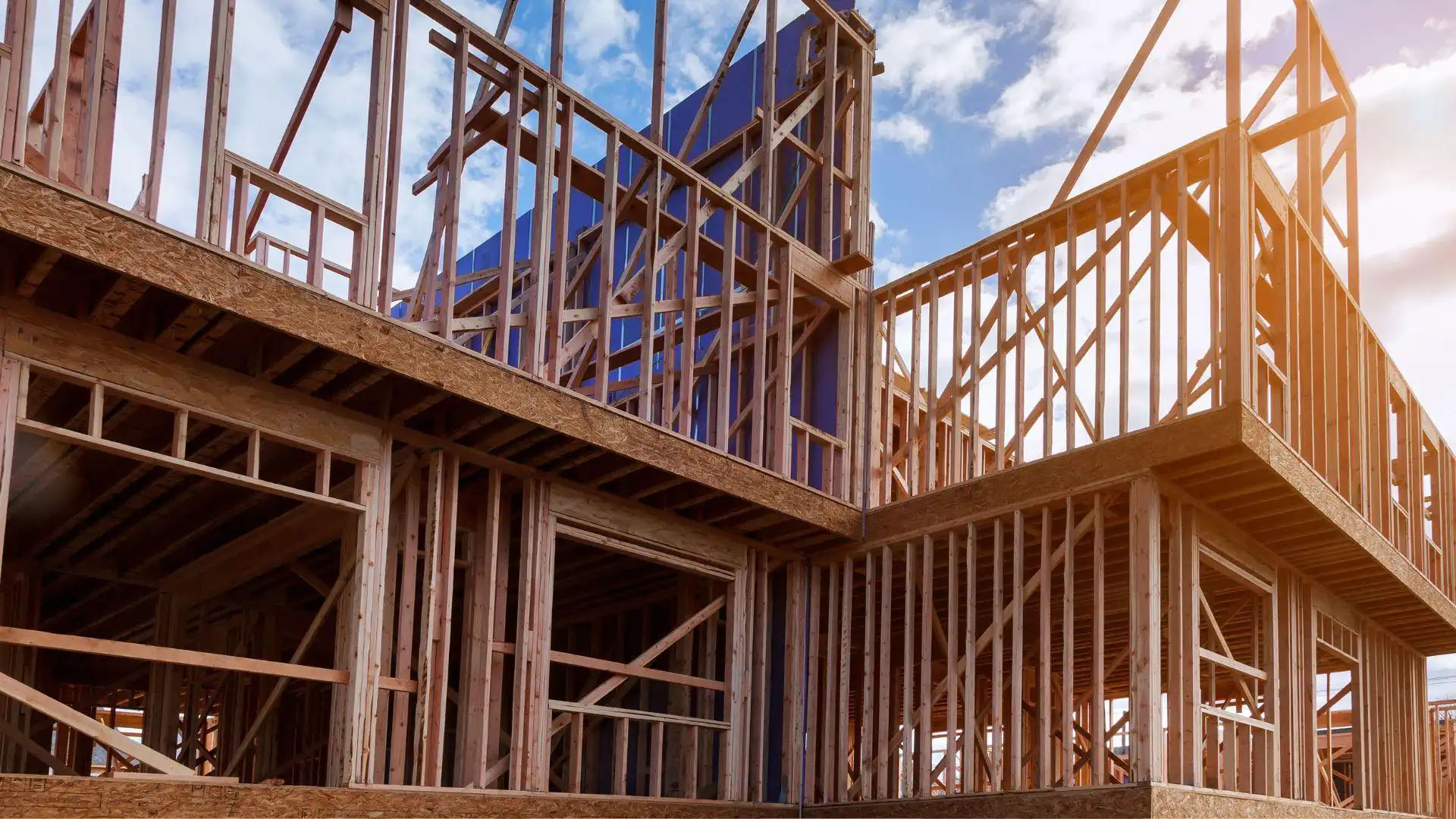 Wooden framework of a multi-level building under construction, with exposed beams and joists beneath a partly cloudy sky.
