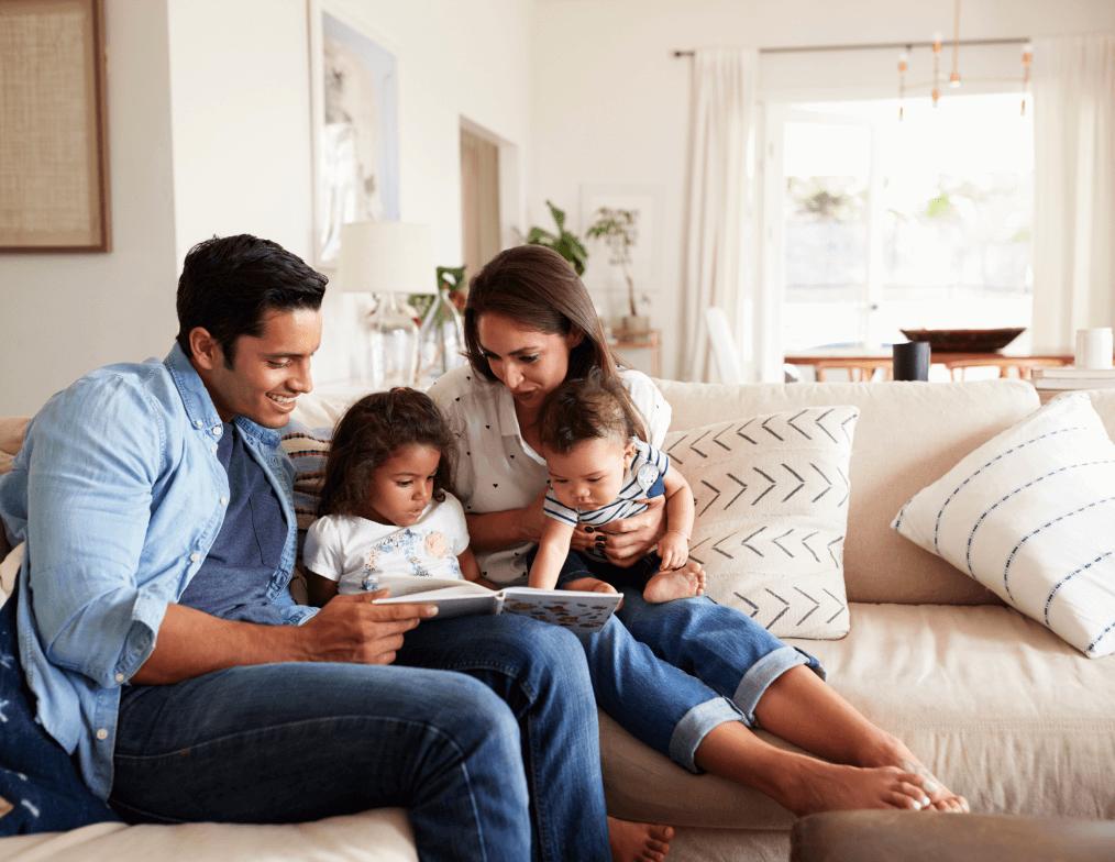 A family of four sits closely together on a beige couch in a bright living room; the father holds an open book, the mother has one child on her lap, and the other child sits between them