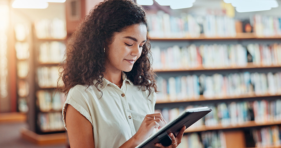 Person standing in a library holding and using a tablet, with bookshelves visible in the background.