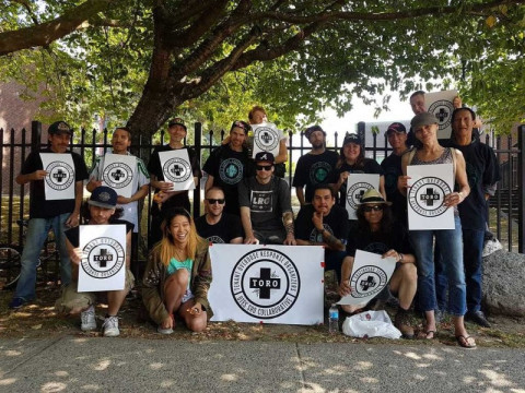 Seventeen people holding white TORO signs stand in front of a black iron fence.
