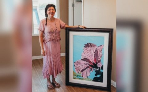 A woman in pink with brown hair stands beside a large, framed painting of a flower.