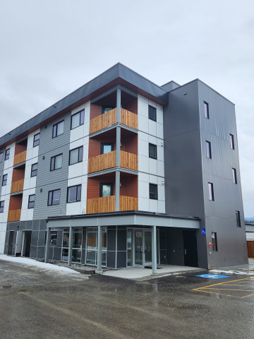 A four-storey grey and white building for seniors, featuring wooden balcony panels and snow around the parking spaces.