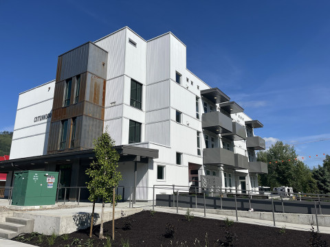 A modern, three-story building with balconies and a flat roof is nestled among trees, with a parking lot in the foreground that includes designated handicap spaces and two parked vehicles—a beige pickup truck and a black car.