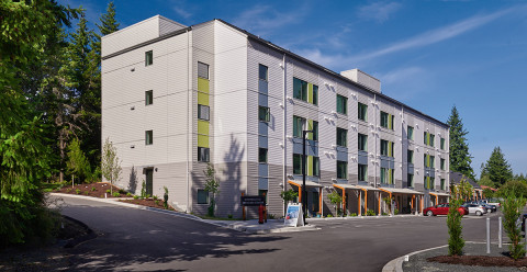 A modern four-story apartment building with light gray siding and green accents, surrounded by trees. The building features large windows, a parking lot with several cars, and a driveway leading to the main entrance.