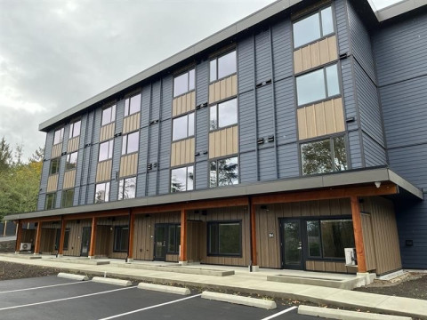 A modern four-story building with wood panelling and dark blue siding, featuring large ground-floor windows and a covered entrance, set beside an empty parking lot.