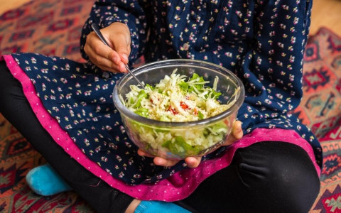 A child sits cross-legged and holds a bowl of greens in their hands.