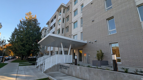 A modern mid-rise apartment building with light gray brick exterior and large windows, featuring a covered entrance with white railings and a concrete sign reading “Heather Place B.” The setting includes a sidewalk, landscaped planters, and tall trees along the street.