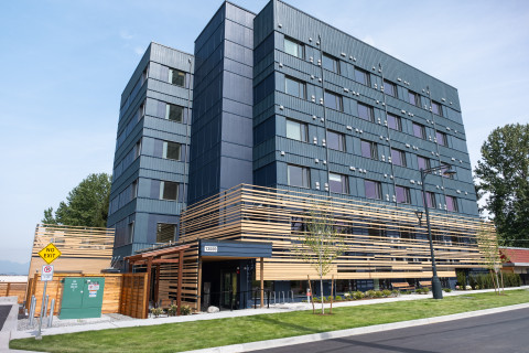 Exterior view of a modern multi‑storey building with dark blue metal siding, horizontal wood slats, large windows, and landscaped grounds.