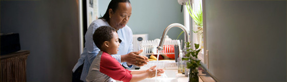 An adult and child standing at a kitchen sink, washing their hands under running water. The countertop holds a dish rack and a small potted plant, with natural light coming through a nearby window.