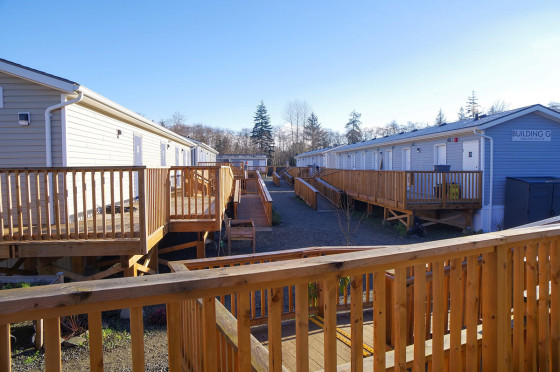 Wooden walkways connect rows of modular housing units under a clear sky, with trees visible in the background.