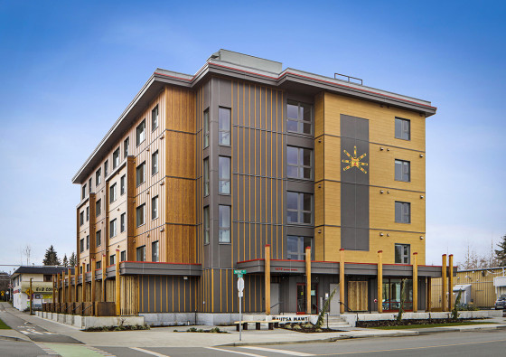 Exterior of Naut'sa mawt, a modern five-storey building with wood and grey panel siding, featuring Indigenous artwork on the facade and located on a street corner in Courtenay, B.C.
