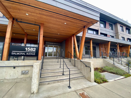 Main entrance of a building with cement stairs and a large wooden roof overhang.