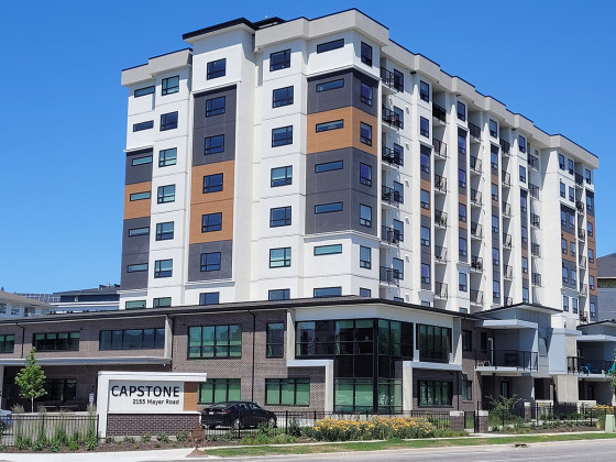 A modern nine-storey apartment building with white, gray, and brown exterior panels, large windows, and balconies, with a sign in front reading "CAPSTONE 2135 Mayer Road" on a clear, sunny day.