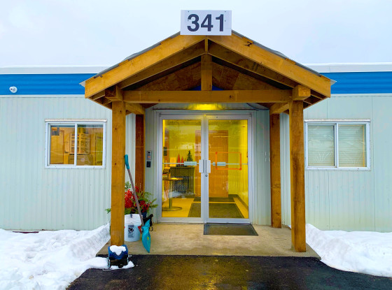 Front entrance of a short white metal-paneled building with blue trim below the roof. A wooden canopy sits above the doors, and the ground is covered in snow.
