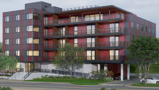 A modern, multi-story apartment building with red and gray exterior walls, elevated on stilts with parking spaces below, and surrounded by trees and greenery; balconies line each floor.
