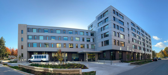 A modern six-storey building with grey and white exterior panels, large windows, and balconies, located at a landscaped street corner under a clear blue sky.
