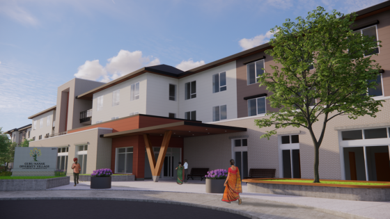 Exterior view of a modern, multi-story building labelled “Guru Nanak Diversity Village” at address 6471, featuring large windows, a wooden-beam-supported covered entrance, landscaped greenery, and several people walking nearby.