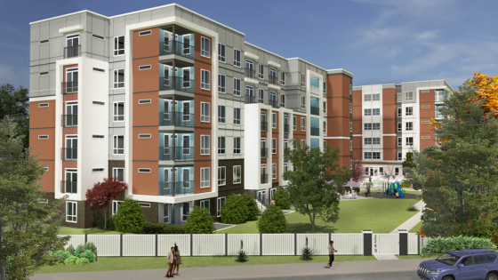 A modern five-storey apartment complex with red brick and white paneling, featuring balconies and large windows. The landscaped courtyard includes green lawns, trees, and a small playground, with a white fence and sidewalk in the foreground.