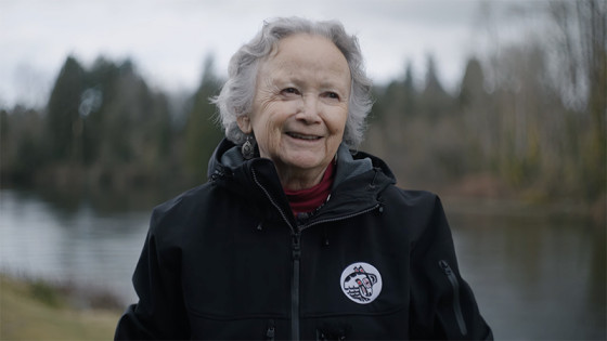 Older adult in a black jacket standing beside a calm lake, with trees and an overcast sky in the background.