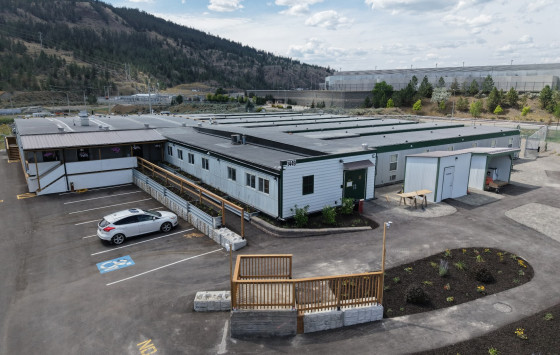 Aerial view of a single-storey modular building complex with accessible ramps, parking areas and landscaped grounds at the base of a tree-covered hillside under a partly cloudy sky.