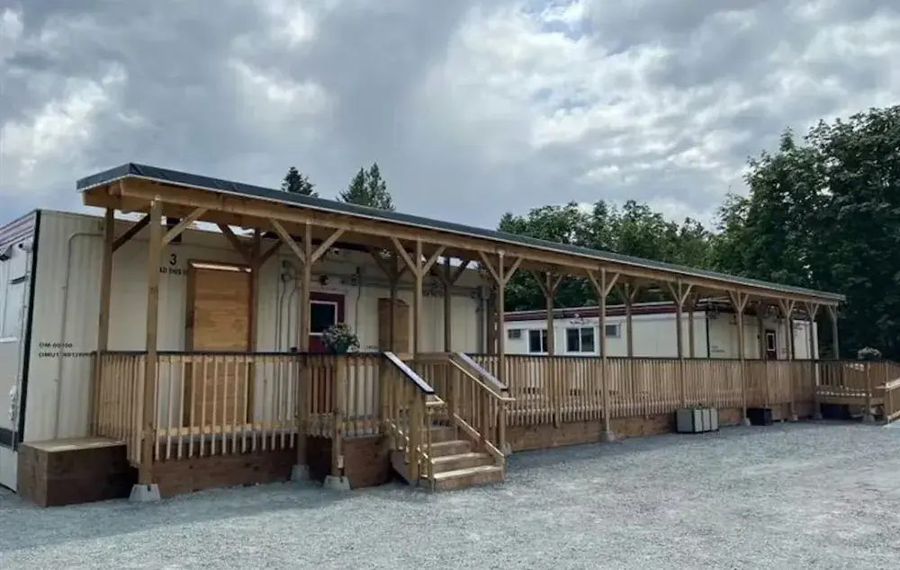Single-storey modular building with a long covered wooden porch, central stairs and ramp, and gravel lot in front, set against trees and a cloudy sky.