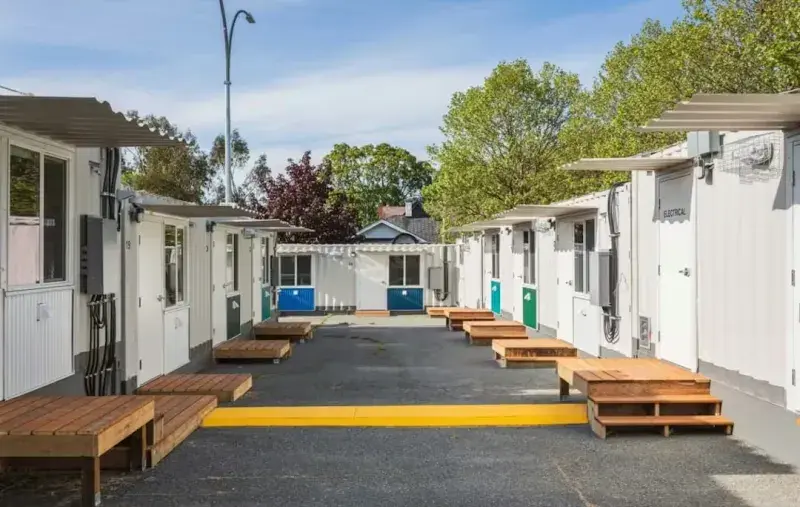 Row of modular housing units facing a central paved walkway, with numbered doors, small wooden steps, a yellow curb marking, and trees in the background.