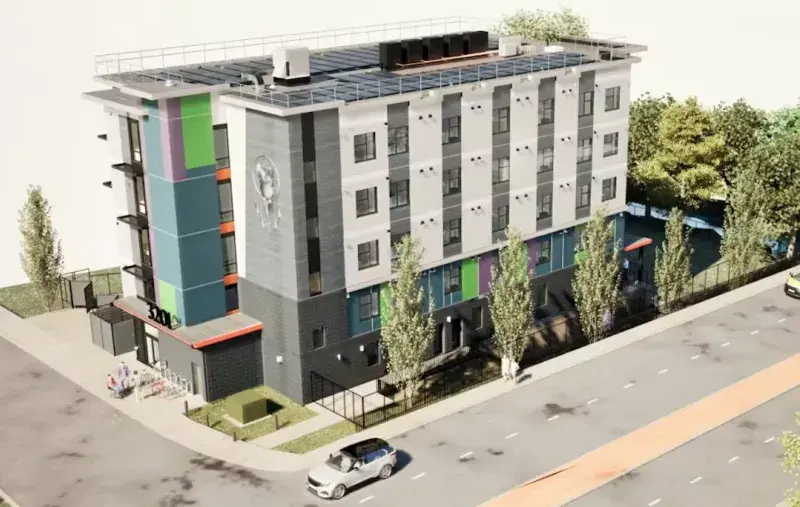 Aerial view of a new five‑storey building with colourful vertical panels, large windows, and a fenced outdoor area, located on a street corner with trees and parked vehicles