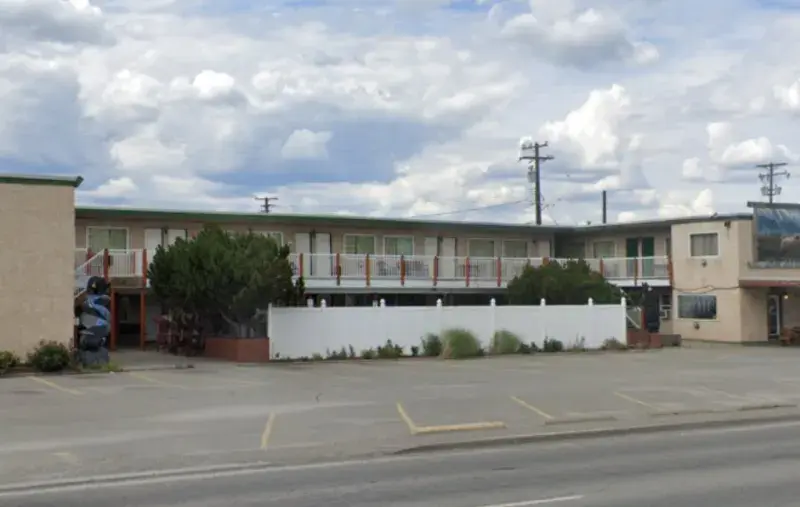 Two-storey beige building with exterior staircases and second-floor walkways, lined with doors and windows, with a white fence and shrubs at the front and an empty parking lot facing a street under a cloudy sky.