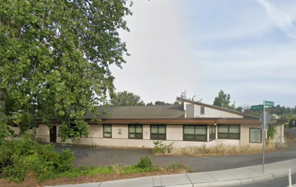 Single-storey beige building with a sloped roof and rows of windows, set back from a sidewalk at a street corner, with large leafy trees and overgrown grass along the front under a partly cloudy sky.