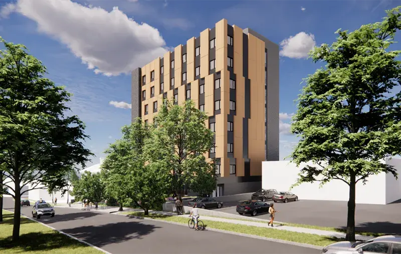 Modern mid-rise building with tan facade on a tree-lined street, with cars and a cyclist under blue sky.